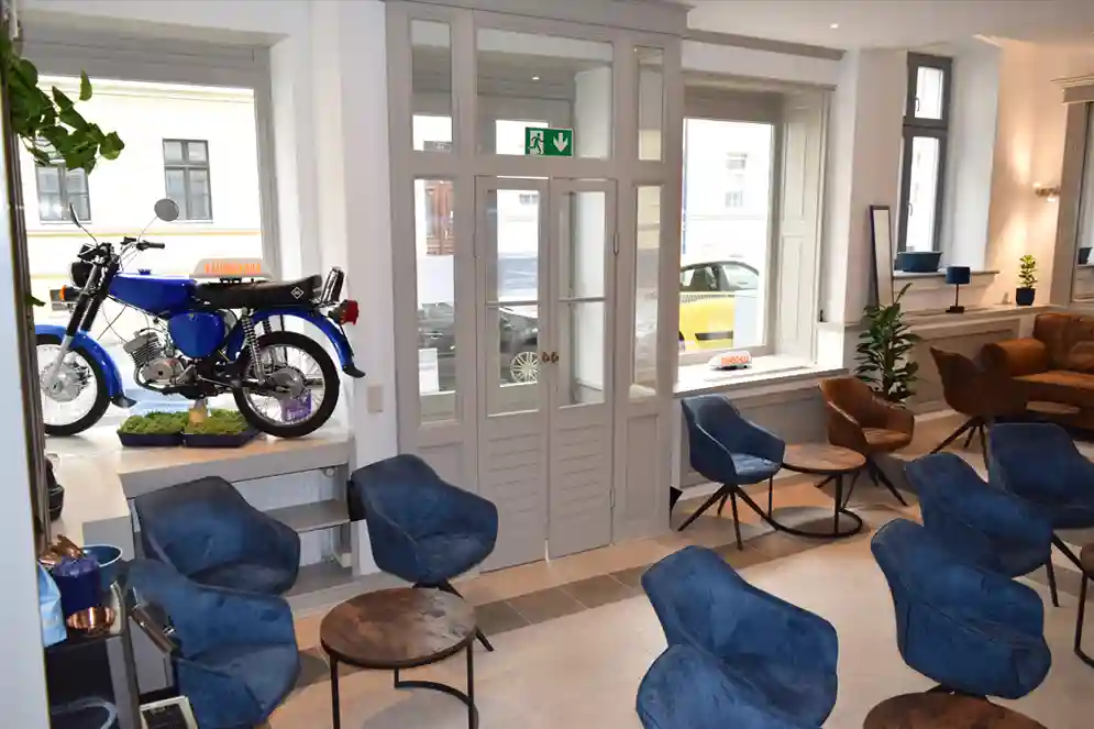 Home 13 Schulungsraum in der Fahrschule "Fahrkompetenz" in Görlitz. Es stehen gemütliche Sessel bereit für den Unterricht in angenehmer Atmosphäre. Ein blaues Simson Moped steht im Schaufenster. Die Doppeleingangstür ist zu erkennen.