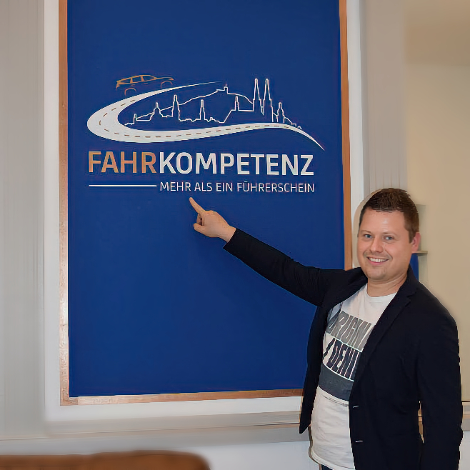 Portrait des Fahrlehrers Philip in der Fahrschule "Fahrkompetenz" in Görlitz. Philip schaut freundlich und zeigt mit dem Finger auf das Logo in seinem Büro.